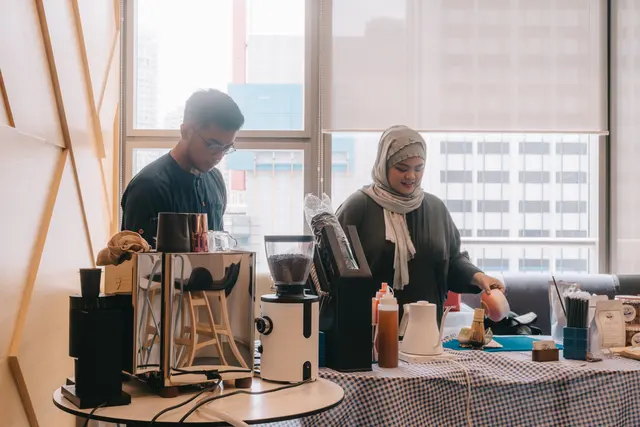 Two Brewprint baristas at a workshop bar by an office window, with a grinder, espresso machine, kettle and tasting cups on the table